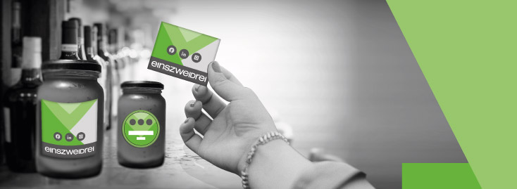 Eine Hand hält eine Karte mit grünen Akzenten und dem Wort „WUNSCHFORMAT“ in fetten Buchstaben vor einer Reihe von Flaschen und Gläsern auf einer Theke; der Hintergrund ist schwarz-weiß, während die Karte und geometrische Formen an der Seite grün gefärbt sind.