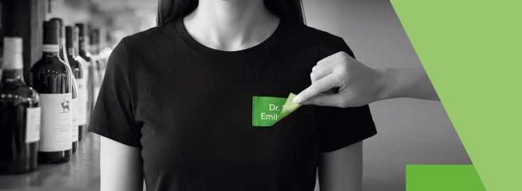 Eine Hand hält eine Karte mit grünen Akzenten und dem Wort „WUNSCHFORMAT“ in fetten Buchstaben vor einer Reihe von Flaschen und Gläsern auf einer Theke; der Hintergrund ist schwarz-weiß, während die Karte und geometrische Formen an der Seite grün gefärbt sind.