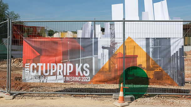 An einem Bauzaun aus Metall ist ein Netzbanner mit abstrakten geometrischen Mustern und teilweise verdecktem weißen Text befestigt; zwei Verkehrskegel stehen vor dem Zaun auf dem Boden.