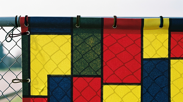 Nahaufnahme eines Maschendrahtzauns mit einer daran befestigten, farbenfrohen Plane, die ein geometrisches Muster aus roten, gelben, blauen und schwarzen Rechtecken aufweist. Die Plane ist mit schwarzen Kabelbindern am Zaun befestigt.