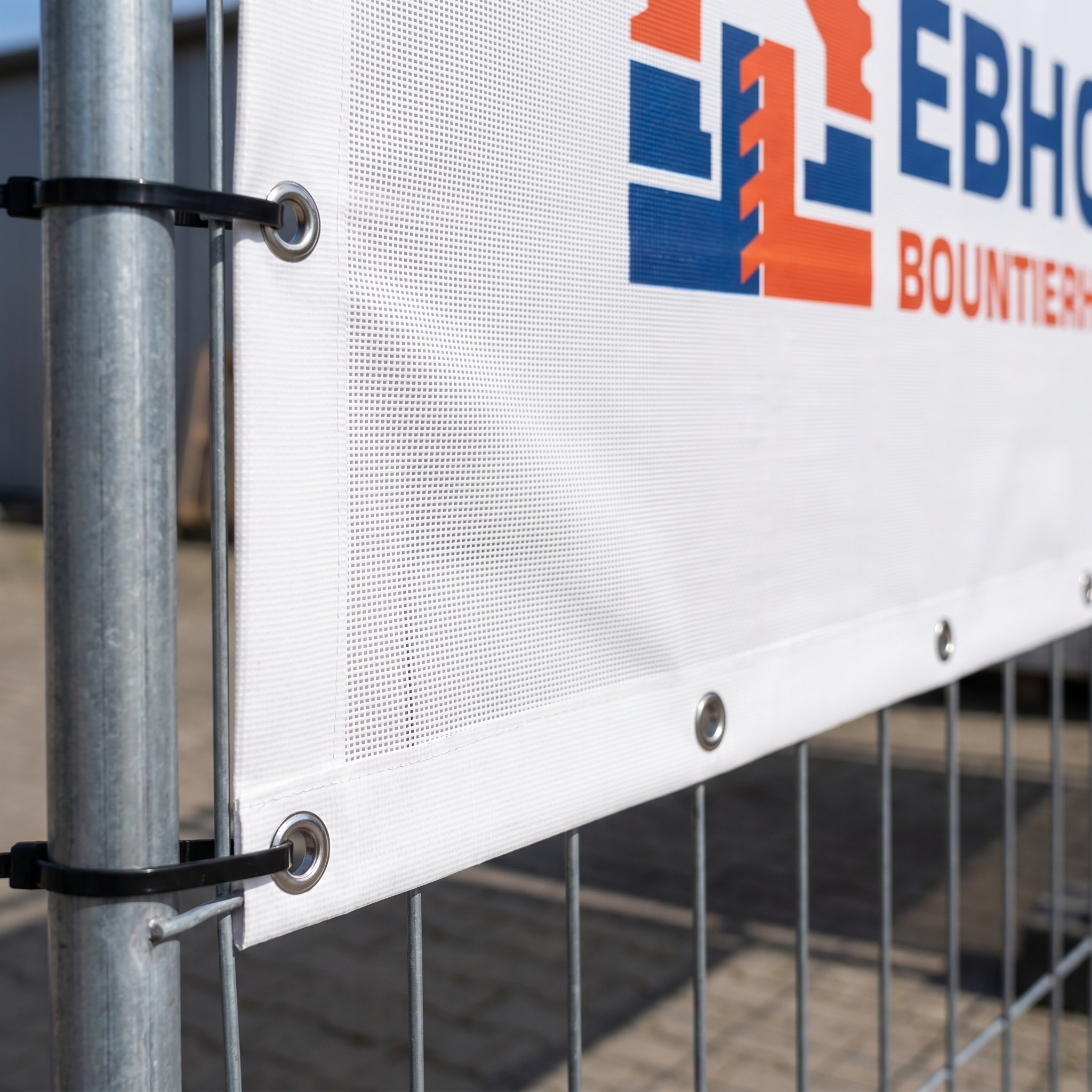 Close-up view of a white mesh banner attached to a metal fence with zip ties, featuring metal grommets and part of a colorful logo and text in blue and orange.