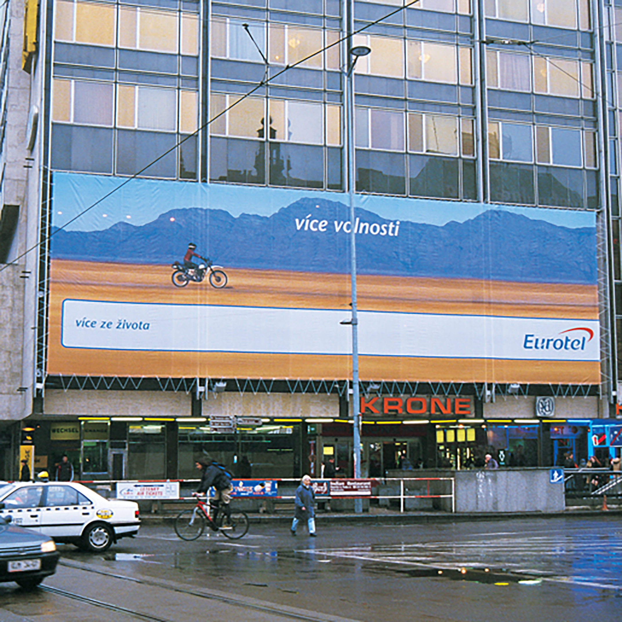 Large outdoor advertisement on a city building shows a person riding a motorcycle against a backdrop of mountains, with the Czech phrases