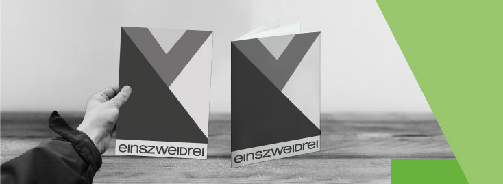 Zwei schwarz-weiße Broschüren mit geometrischen Mustern und dem Text „einszweidrei“ auf dem Umschlag, eine in der Hand gehalten, die andere auf einer Holzfläche stehend, mit einem grünen grafischen Element im Hintergrund.