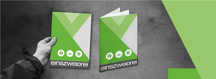 Eine Hand hält eine grün-weiße Broschüre mit geometrischen Mustern. Daneben liegt eine geöffnete Version derselben Broschüre, deren gefaltetes Inneres sichtbar ist. Die Broschüren enthalten Symbole für Facebook, LinkedIn und Instagram sowie den Schriftzug „einszweidrei“ am unteren Rand, alles vor einem strukturierten grau-grünen Hintergrund.