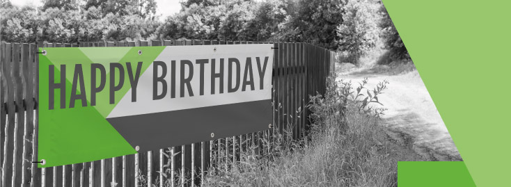 An einem Holzzaun entlang eines Feldwegs ist ein großes PVC-Banner mit der Aufschrift „HAPPY BIRTHDAY“ in fetten Buchstaben befestigt. Das Banner ist mit grünen und grauen geometrischen Mustern und grünen Akzenten versehen.