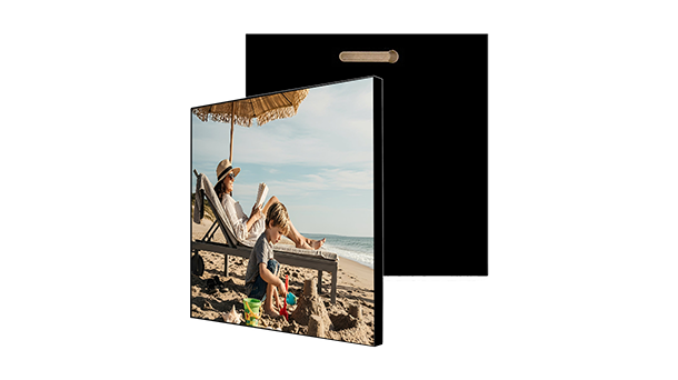 Quadratische MDF-Wandplatte mit einem aufgedruckten Foto einer Frau, die auf einem Strandstuhl unter einem Strohschirm entspannt, während vor ihr ein Kind im Sand spielt; die Platte hat eine schwarze Rückseite und einen Aufhängeschlitz.