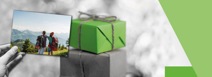 Eine Hand hält ein farbenfrohes Foto von zwei Wanderern, die eine Berglandschaft betrachten. Vor dem Foto stehen zwei gestapelte Geschenke, eines davon in leuchtend grünes Papier gewickelt, mit monochromem Hintergrund und grünen Akzenten.