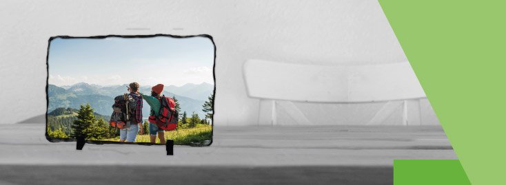 Ein weißes Kissen mit einem aufgedruckten Foto von zwei Personen mit Rucksäcken, die im Freien stehen und eine Berglandschaft betrachten, vor einem grauen Hintergrund mit grünen geometrischen Akzenten.