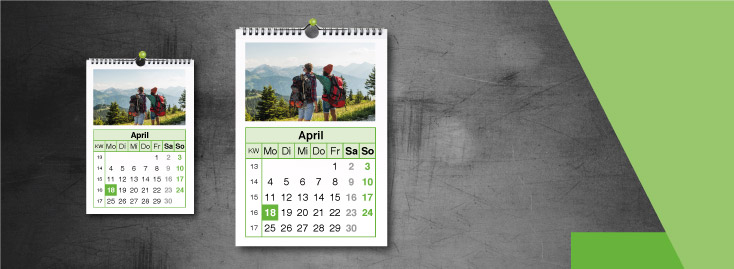 Vor einem grauen Hintergrund sind zwei Wandkalender für April angebracht, die jeweils ein Foto von zwei Wanderern mit Rucksäcken zeigen, die auf Berge und Bäume blicken. Das darunter liegende Kalenderraster zeigt die Tage des Monats an.