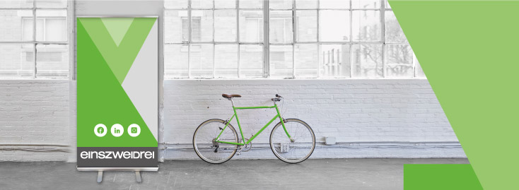 Ein grün-weißes Roll-Up-Banner mit Social-Media-Symbolen und dem Text „einszweidrei“ steht neben einem grünen Fahrrad vor einer weißen Backsteinmauer und großen Industriefenstern.