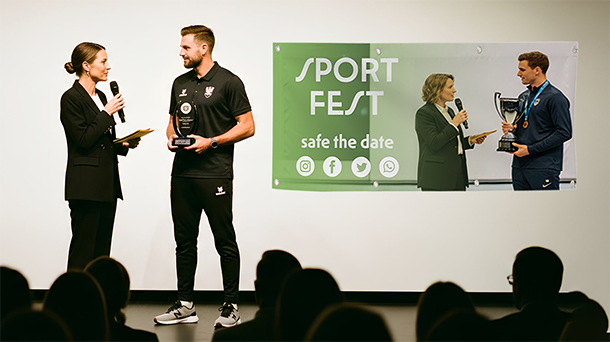 Eine Frau und ein Mann stehen auf der Bühne. Die Frau hält ein Mikrofon, der Mann einen Pokal. Zuschauer beobachten das Geschehen. Hinter ihnen hängt ein grün-weißes Banner mit der Aufschrift „SPORT FEST, Termin vormerken“, Social-Media-Symbolen und einem Foto, das eine Frau zeigt, die einem Mann einen Pokal überreicht.