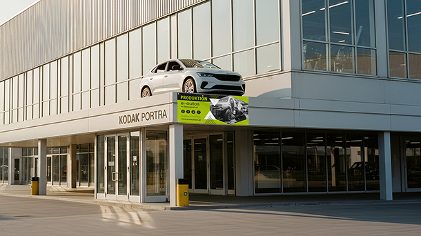 Ein modernes Geschäftsgebäude mit großen Glasfenstern, einem weißen Auto auf dem oberen Sims und einem leuchtend grünen PVC-Banner mit der Aufschrift „Produktion“, das von der Fassade hängt.
