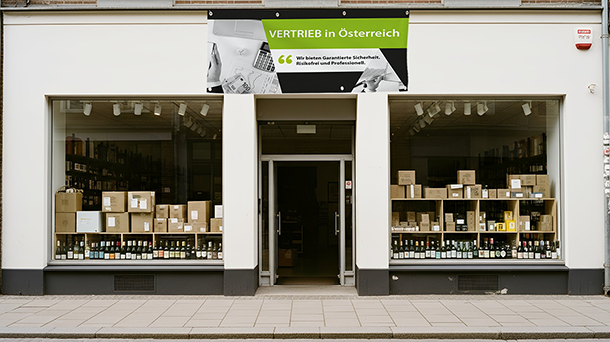 Ein Schaufenster mit großen Schaufenstern, in denen Regale und Stapel verschiedener verpackter Produkte zu sehen sind, über dem Eingang ein auffälliges grün-schwarzes PVC-Banner mit der Aufschrift „VERTRIEB in Österreich“ und darunter ein weiterer deutscher Text über garantierte Sicherheit, Zuverlässigkeit und Professionalität.
