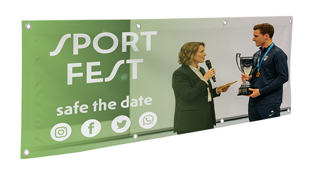 Ein grünes PVC-Banner mit der Aufschrift „Sportfest“ und dem Text „Termin vormerken“ sowie Social-Media-Symbolen ist zu sehen. Davor interviewt eine Frau mit Mikrofon einen Mann, der einen Pokal hält und eine Medaille trägt.