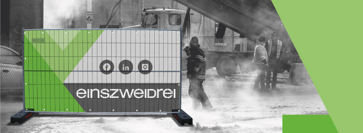 Eine Baustelle mit mehreren Arbeitern wird in Schwarzweiß gezeigt, während im Vordergrund ein farbenfrohes grün-graues Maschendrahtzaunbanner den Text „einszweidrei“ und Social-Media-Symbole anzeigt.