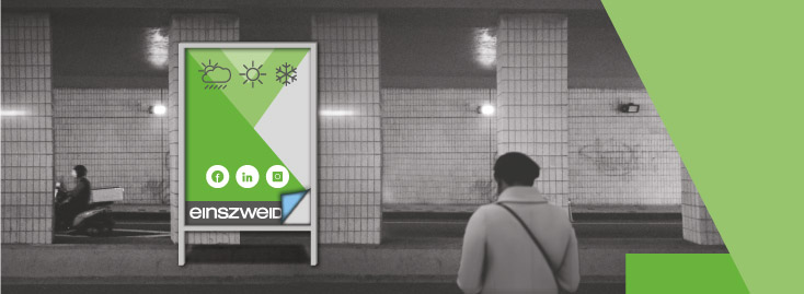 Eine Schwarz-Weiß-Aufnahme einer U-Bahn-Szene zeigt zwei Personen, in deren Mitte ein auffälliges grün-weißes Immobilienplakat prangt, auf dem Wettersymbole, Logos sozialer Medien und das Wort „einszweid“ zu sehen sind; eine große grüne geometrische Form akzentuiert die rechte Seite des Bildes.