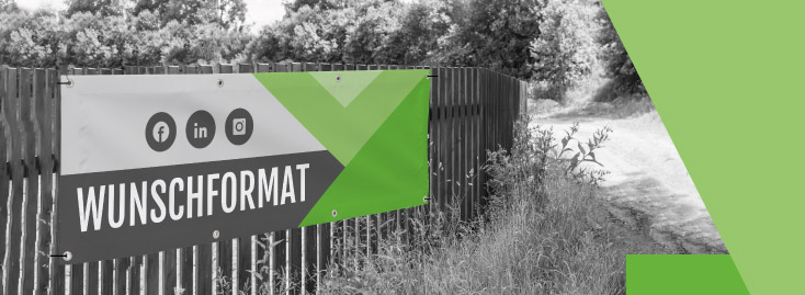 Ein grün-weißes PVC-Banner mit der Aufschrift „WUNSCHFORMAT“ hängt an einem schwarzen Zaun neben einem Feldweg. Im Hintergrund sind teilweise entsättigte Bäume und Sträucher zu sehen, überlagert von grünen Grafikelementen. Auf dem Banner sind Social-Media-Symbole für Facebook, LinkedIn und Instagram abgebildet.