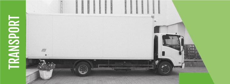 Neben einem Gebäude steht ein weißer Kastenwagen. Links davon prangt auf einem grünen Schild senkrecht der Schriftzug „TRANSPORT“. Das Bild deutet auf den Transport von Druckerzeugnissen hin.