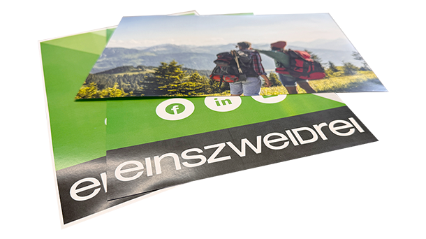Ein Stapel Werbeflyer, wobei der oberste zwei Wanderer mit Rucksäcken zeigt, die auf eine malerische Berglandschaft blicken, und der darunter grüne Social-Media-Symbole und den Text „einszweidrei“ auf schwarzem Hintergrund aufweist.
