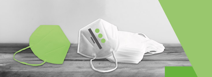 Auf einer Holzoberfläche ist ein Stapel weißer FFP2-Masken ausgestellt. Eine weiße Maske zeigt grüne und graue grafische Elemente, links daneben befindet sich eine separate grüne Maske. Der Hintergrund ist überwiegend neutral, grüne Formen rahmen das Bild ein.