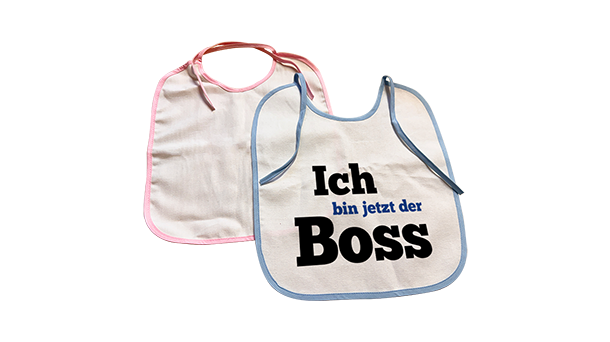 Es werden zwei Baby-Lätzchen gezeigt; eines hat einen rosa Rand, das andere einen blauen Rand mit dem deutschen Text „Ich bin jetzt der Boss“ in fetten Buchstaben.
